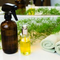 Amber and clear glass spray bottles. Rolled green towels in a spa setting. Green plant decor in background. Bathroom white countertop.