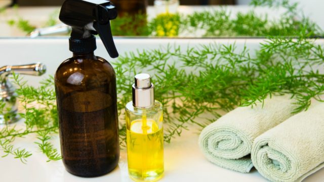 Amber and clear glass spray bottles. Rolled green towels in a spa setting. Green plant decor in background. Bathroom white countertop.