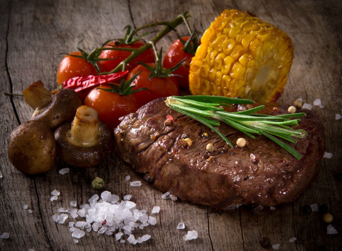 Cooked steak with tomatoes, corn, and mushroom on a rustic wood background