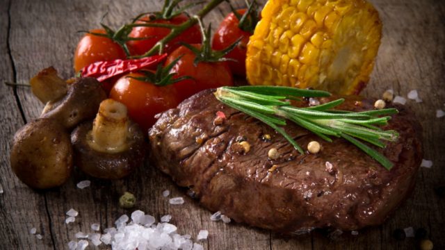 Cooked steak with tomatoes, corn, and mushroom on a rustic wood background